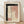 May include: Framed abstract watercolor artwork leaning against white wall on light wood floor. The piece features soft pink rectangular shapes, bold black brushstrokes forming an L-shape, and a vertical line of small black dots on cream textured paper. Natural light casts diagonal shadows across the scene. Light wood frame complements the minimal, modern aesthetic.