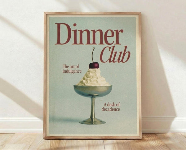 May include: Framed vintage-style poster with sage green background featuring red text reading "Dinner Club" and "The art of indulgence" alongside "A dash of decadence." Center shows retro illustration of glass coupe filled with white whipped cream topped with dark cherry. Natural wood frame sits on light wooden floor against white wall with soft window light shadows.