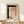 May include: Abstract watercolor artwork in wooden frame displayed on light wood shelf against cream wall. Painting features black brushstrokes forming an L-shape with terracotta pink washes and dotted line down center on textured paper. Small wrapped white gift box with pine sprig sits in front of frame. Two hands rest on shelf edges.