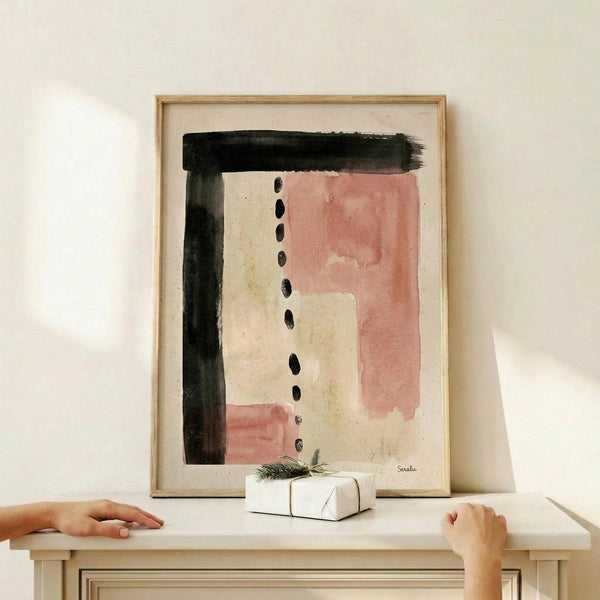 May include: Abstract watercolor artwork in wooden frame displayed on light wood shelf against cream wall. Painting features black brushstrokes forming an L-shape with terracotta pink washes and dotted line down center on textured paper. Small wrapped white gift box with pine sprig sits in front of frame. Two hands rest on shelf edges.