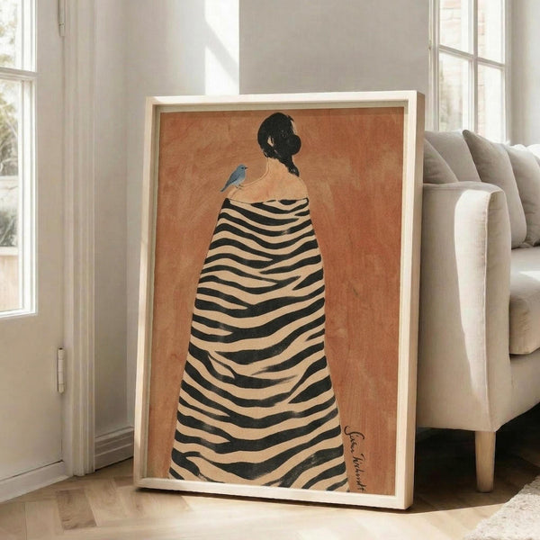 May include: Framed artwork leaning against white wall next to beige sofa in bright living room. The poster shows a woman from behind wearing a black and white striped zebra-patterned cloak or dress against an orange-brown background, with a small blue bird perched on her shoulder. Her dark hair is styled in a low bun. The piece has a minimalist, contemporary art style in a light wood frame.