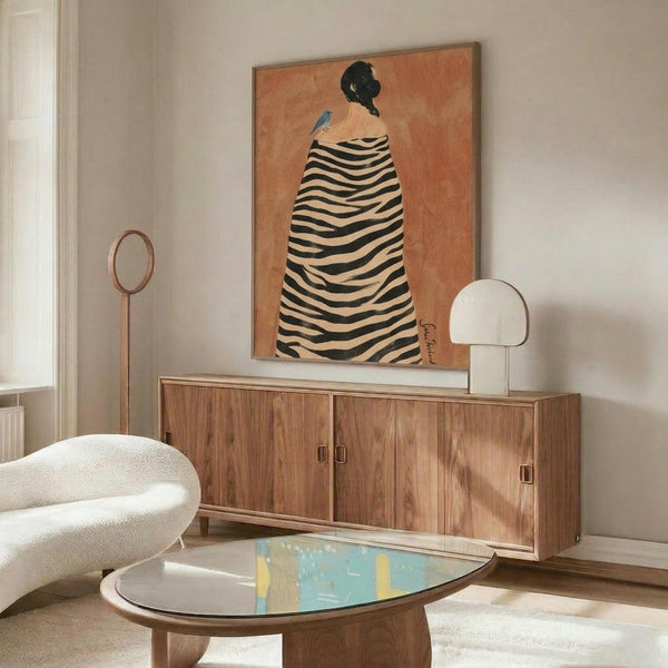 May include: Contemporary living room interior featuring a framed art print on white wall above wooden sideboard. The artwork shows a figure in black and white zebra-striped cloak with small blue bird, set against warm orange background. Room has cream curved chair, white mushroom lamp, oval glass coffee table, and natural lighting creating soft shadows.