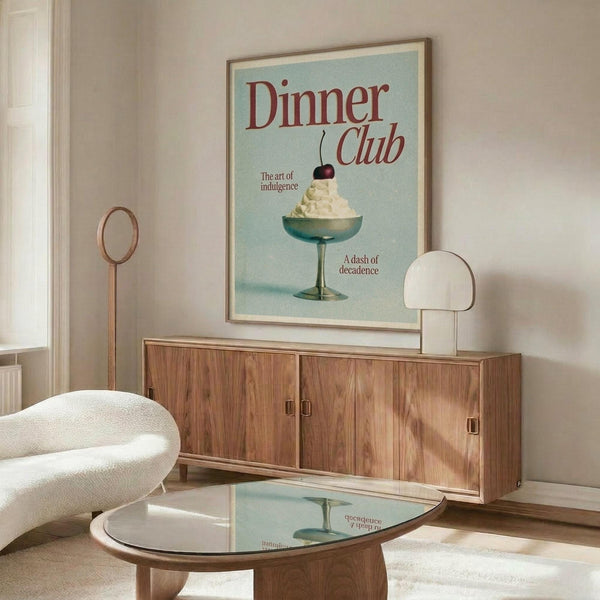 May include: Framed vintage-style poster with light blue background showing dessert in green glass bowl topped with whipped cream and cherry, red text reading "Dinner Club" and "The art of indulgence" and "A dash of decadence". Displayed above walnut wood credenza in modern living room with white sculptural chair, round glass coffee table, brass floor lamp and table mirror.