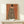 May include: Framed art print displayed on light wood console table against white wall. Print features woman in black and white striped zebra-patterned cloak on terracotta orange background, with small blue bird perched on her shoulder. Natural wood frame, small white wrapped gift with greenery sits in front of artwork.