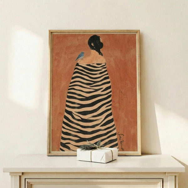 May include: Framed art print displayed on light wood console table against white wall. Print features woman in black and white striped zebra-patterned cloak on terracotta orange background, with small blue bird perched on her shoulder. Natural wood frame, small white wrapped gift with greenery sits in front of artwork.