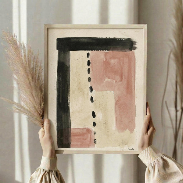 May include: Person holding framed abstract art print with light wood frame. Artwork features dusty pink watercolor rectangles, dark green brushstrokes forming an L-shape, and vertical line of black oval dots on cream textured paper. Person wears beige ruffle-sleeve top, holds dried wheat stems in one hand. Soft natural lighting against neutral curtained background.