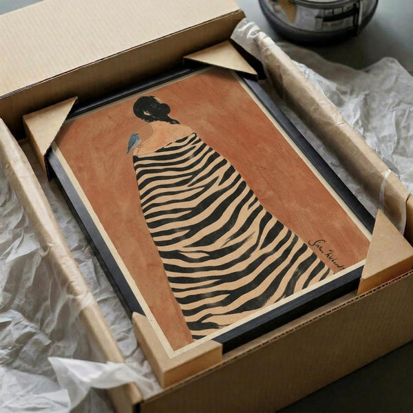 May include: Framed art print featuring a figure in profile wearing a black and cream striped zebra-patterned cloak against a warm terracotta background. The minimalist illustration shows the person with dark hair in an updo. The black-framed print is carefully packaged in a brown cardboard box with protective white tissue paper, displayed on a white surface alongside wooden tubes and packaging materials.