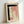 May include: Framed abstract artwork leaning against white wall, featuring bold black brushstrokes forming an L-shape, soft pink watercolor washes, and vertical line of black dots on cream paper, displayed in light wood frame with natural lighting casting gentle shadows