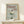 May include: Framed vintage-style poster with sage green background featuring elegant cocktail glass filled with white cream or ice cream topped with dark cherry. Red text reads "Dinner Club" with "The art of indulgence" and "A dash of decadence" in smaller text. Natural wood frame sits on light wooden surface next to small white wrapped gift with evergreen sprig against cream wall.