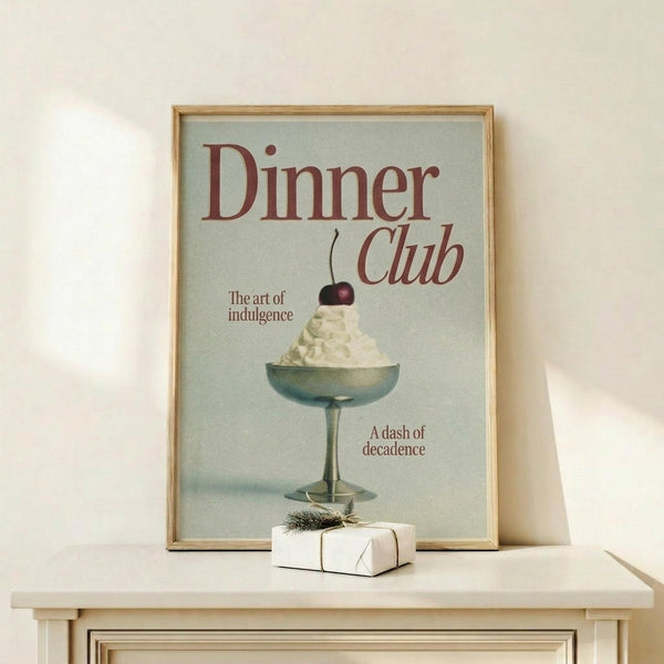 May include: Framed vintage-style poster with sage green background featuring elegant cocktail glass filled with white cream or ice cream topped with dark cherry. Red text reads "Dinner Club" with "The art of indulgence" and "A dash of decadence" in smaller text. Natural wood frame sits on light wooden surface next to small white wrapped gift with evergreen sprig against cream wall.