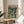 May include: Framed poster featuring anime characters in an urban nighttime scene with neon lighting and Japanese text at top, displayed on a light wood round side table next to a beige upholstered chair with white throw pillow, natural lighting casting shadows on cream wall with plant leaves visible in corner.