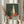 May include: Person holding framed still life painting featuring green round vase, tall cream bottle, small red vase, and red apple arranged on white cloth against dark green background. Artwork displayed in thin light wood frame. Person wearing beige ruffled sleeves holds dried wheat stalks, standing by white curtained window in soft natural lighting.