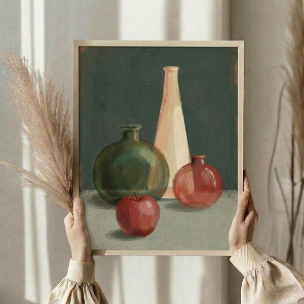 May include: Person holding framed still life painting featuring green round vase, tall cream bottle, small red vase, and red apple arranged on white cloth against dark green background. Artwork displayed in thin light wood frame. Person wearing beige ruffled sleeves holds dried wheat stalks, standing by white curtained window in soft natural lighting.