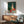 May include: Still life painting in wooden frame showing green and white ceramic vases with red apple and small red vessel on light surface against teal background, displayed above mid-century modern wooden credenza with white table lamp, next to sage green upholstered armchair with wooden arms in sunlit room with white walls