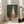 May include: Framed still life painting leaning against beige sofa in bright living room with hardwood floors and large windows. Artwork features red apple and three glass bottles - green, cream, and orange-red - against teal background with gray surface. Natural wood frame complements neutral interior decor.