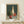 May include: Framed still life painting displayed on light wood surface showing green glass bottle, tall cream vase, small red glass vessel, and red apple against dark teal background. Natural lighting creates soft shadows on white wall. Small wrapped gift with evergreen sprig sits beside the light wood frame.