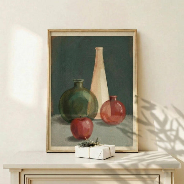 May include: Framed still life painting displayed on light wood surface showing green glass bottle, tall cream vase, small red glass vessel, and red apple against dark teal background. Natural lighting creates soft shadows on white wall. Small wrapped gift with evergreen sprig sits beside the light wood frame.