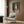 May include: Still life poster print featuring bottles and vases in green, cream, and coral tones with red apples on teal background, displayed in light wood frame on brown wood paneled wall above beige upholstered armchair with geometric patterned pillow, next to brass floor lamp and dark round side table with open book.