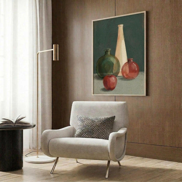 May include: Still life poster print featuring bottles and vases in green, cream, and coral tones with red apples on teal background, displayed in light wood frame on brown wood paneled wall above beige upholstered armchair with geometric patterned pillow, next to brass floor lamp and dark round side table with open book.