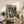 May include: Framed anime poster featuring orange and purple mecha robot standing beside young person in white and teal outfit, with Japanese text header, displayed on rustic wooden sideboard next to fern plant, books, and ceramic vase in cozy living room setting with beige armchair visible.