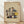 May include: Framed modernist architectural drawing showing an abstract geometric building composition with colorful rectangular modules in yellow, blue, red, and gray stacked at various angles. The artwork features clean lines and appears to be a vintage mid-century design study rendered on textured beige paper, displayed in a natural wood frame against white walls on light wooden flooring with soft natural lighting.