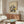 May include: Abstract architectural artwork with colorful geometric building forms in yellow, blue, red and gray tones, displayed in light wood frame above walnut sideboard. Modern living room setting with cream upholstered chair, oval glass coffee table, and white table lamp. Neutral walls with natural lighting from window.