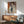 May include: Framed modernist portrait artwork hanging above wood credenza in mid-century modern room setting. Abstract portrait features figure with dark hair and geometric clothing in muted tones of beige, orange, and navy. Natural wood frame complements teal upholstered chair and ceramic table lamp. Warm sunlight creates diagonal shadows across white wall.