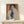 May include: Framed modernist portrait print displayed on light wooden shelf against white wall. Artwork features stylized figure with asymmetrical face in muted tones of purple, orange, and navy blue, rendered in geometric cubist style. Natural wood frame complements minimal setting with small wrapped gift box nearby.