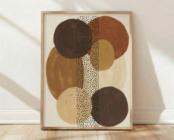 May include: Framed abstract art print featuring overlapping circles in earthy brown, rust, golden yellow and cream tones with black polka dot pattern running vertically through center, displayed in light wood frame leaning against white wall on wooden floor with natural window light creating soft shadows