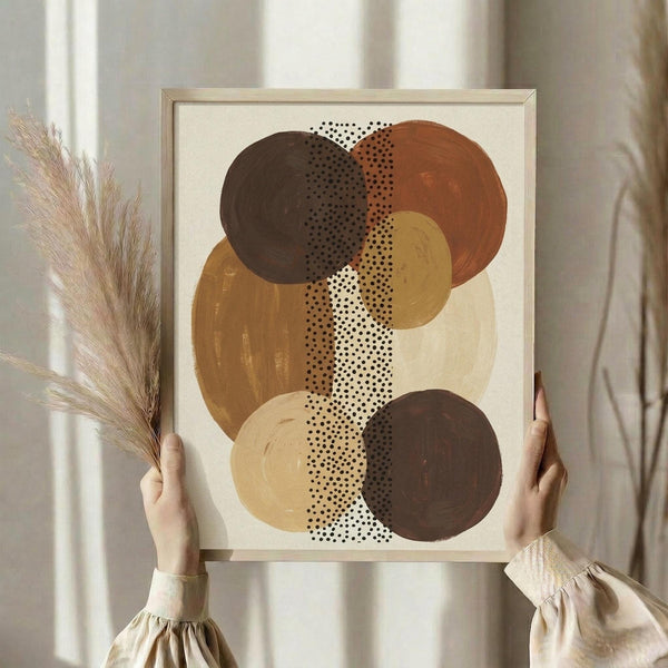 May include: Person holding framed abstract art print featuring overlapping circles in earthy tones of brown, rust orange, mustard yellow, and cream with dotted patterns running vertically through the composition. Light wood frame against neutral wall with dried pampas grass visible to the side.