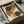 Abstract art print with overlapping circles in warm earth tones including rust, brown, tan, and cream colors. Features dotted pattern details running vertically through the composition. Print is displayed in a black frame inside an open cardboard shipping box with protective tissue paper and wooden corner guards, sitting on white fabric with a dark glass object visible in the background.