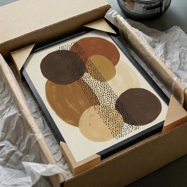 Abstract art print with overlapping circles in warm earth tones including rust, brown, tan, and cream colors. Features dotted pattern details running vertically through the composition. Print is displayed in a black frame inside an open cardboard shipping box with protective tissue paper and wooden corner guards, sitting on white fabric with a dark glass object visible in the background.