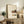 May include: Framed Japanese calligraphy poster with kitchen still life illustrations displayed on rustic wooden sideboard. Cream-colored artwork features black text, coffee pot, teacup, fruit, and kitchen items in soft watercolor style. Natural wood frame complements surrounding decor including fern plant, ceramic vase, books, and beige upholstered furniture in warm, minimalist living space with white walls.
