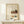 May include: Framed Japanese poster with cream background featuring kitchen still life illustrations - teapot, plants, pastries and teacup in warm earth tones. Black Japanese calligraphy text on left side. Natural wood frame displayed on light wooden surface next to small white wrapped gift with evergreen sprig. Soft lighting creates gentle shadows on neutral wall background.