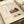 May include: A framed poster with Japanese calligraphy and watercolor illustrations showing kitchen items on wooden surfaces - a dark teapot, white bowl with green plant, oranges on a plate, and a teal cup. The artwork has cream background with black vertical text on the left side and smaller text at bottom. The frame appears light wood-toned, displayed on white bedding with soft natural lighting.