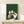 May include: Framed poster print on light wood shelf showing five cats gathered around wooden table playing board game on forest green textured background. Cats in various colors - white, gray, black, and cream - sit contentedly around chess or checkers board. Small wrapped gift package with dried botanicals sits beside frame on natural wood surface against cream wall.