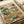 May include: Vintage-style poster print displayed on white paper showing two orange tabby cats in sunglasses and Hawaiian shirts sitting at a wooden table on a tropical beach. The cats hold margaritas while surrounded by Mexican food including tacos, guacamole, chips, and shrimp. Palm trees and blue ocean create the background under a pink sunset sky. The poster has a weathered, retro aesthetic with "FELINE FIESTA" text at the top in colorful letters.