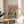May include: Framed poster displaying two cartoon penguins at a party table with colorful "PENGUIN PARTY" text above them. One penguin wears a tropical shirt and party hat, the other has sunglasses and a bow tie. Table shows pizza, cookies, fish, drinks and champagne bottle against coral pink background. Wooden frame sits on round wooden stool next to beige chair and plant in bright, sunlit room.