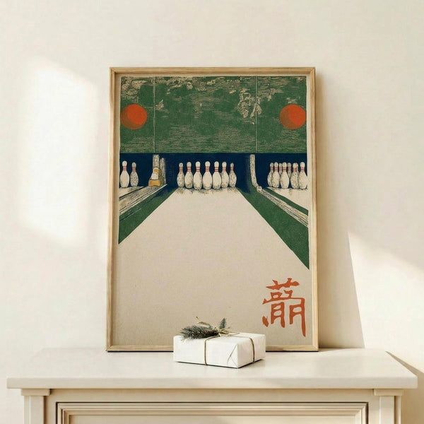 May include: Framed vintage Japanese art print depicting a bowling alley interior with white pins, green lanes, and red circular lights. The artwork features Japanese characters in orange text and has a woodblock print aesthetic. Displayed on a light wood console table with a small wrapped gift box beside it against a cream wall.