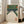 May include: Framed vintage Japanese art print depicting a bowling alley scene with green walls, cream-colored lanes, white bowling pins, and red circular elements at the top. Orange Japanese characters appear in the bottom right corner. The print is displayed in a light wood frame leaning against a white wall next to a beige upholstered chair in a bright, modern living room with large windows.