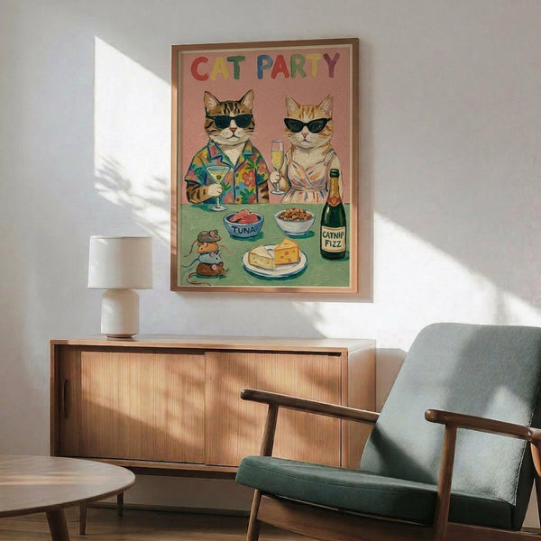 May include: Framed poster showing two cats wearing sunglasses at a party table with colorful "CAT PARTY" text above them. The tabby cat wears a tropical shirt and holds a martini, while the orange cat in a dress holds champagne. Table has tuna bowls, cheese, toy mice, and a "Catnip Fizz" bottle on pink and green background. Displayed above a mid-century modern wood credenza with white table lamp, next to a teal upholstered armchair in a bright living room.