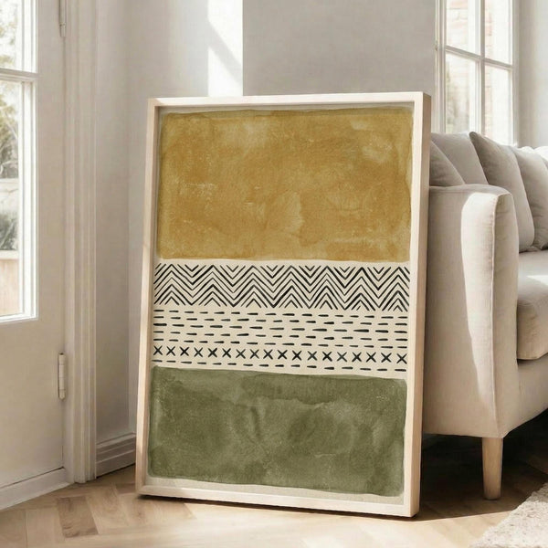 May include: Large framed abstract art print leaning against white wall next to beige sofa in bright living room. Artwork features horizontal bands of ochre yellow and olive green watercolor sections separated by black and white geometric patterns including chevrons, dashes, and crosses on cream background in light wood frame.