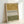 Abstract artwork featuring horizontal bands of muted ochre yellow and olive green watercolor sections separated by a white middle band with black geometric patterns including chevron zigzags, dashed lines, and small x marks. The piece is displayed in a light natural wood frame leaning against a white wall, creating soft shadows.