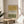 May include: Modern abstract art print in wooden frame displaying mustard yellow and olive green watercolor blocks separated by black and white geometric tribal patterns including zigzags, dashes, and crosses. Frame sits on round wooden side table next to beige upholstered chair with white pillow and plant leaves visible in bright, minimalist living room setting.
