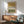 May include: Abstract art print with ochre yellow and olive green watercolor blocks separated by black and white geometric tribal patterns, displayed in light wood frame above mid-century modern credenza. Sage green upholstered chair with wooden arms visible in foreground, white ceramic table lamp, warm sunlight casting shadows on white wall.