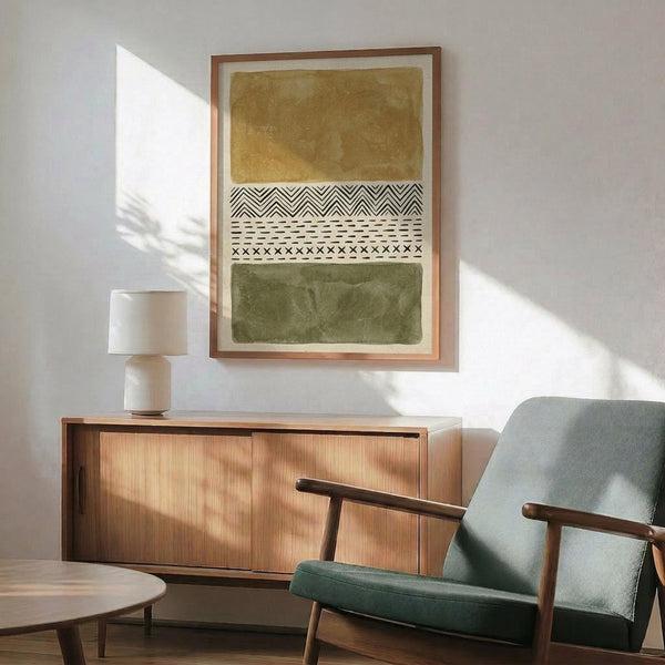 May include: Abstract art print with ochre yellow and olive green watercolor blocks separated by black and white geometric tribal patterns, displayed in light wood frame above mid-century modern credenza. Sage green upholstered chair with wooden arms visible in foreground, white ceramic table lamp, warm sunlight casting shadows on white wall.