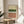 May include: Abstract geometric art print in wooden frame on round wooden side table next to beige upholstered chair. Artwork features horizontal bands of cream, green watercolor wash, white and terracotta sections with black polka dot patterns. Natural lighting creates soft shadows on neutral wall. Plant leaves visible in corner.