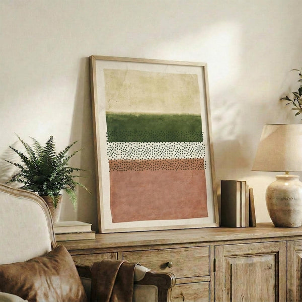 May include: Abstract art print with horizontal bands in cream, forest green, white with black dots, and terracotta pink, displayed in light wood frame on rustic wooden sideboard alongside fern plant, books, and ceramic vase in cozy living room setting with leather armchair