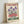 A vintage-style poster in a light wooden frame leaning against a white wall. The cream-colored print features purple irises and red flowers with green stems and leaves in a folk art illustration style. "FLOWER MARKET" appears in burgundy letters at the top, with "AMSTERDAM" in green letters at the bottom. The artwork has a weathered, textured appearance with subtle distressing around the edges.