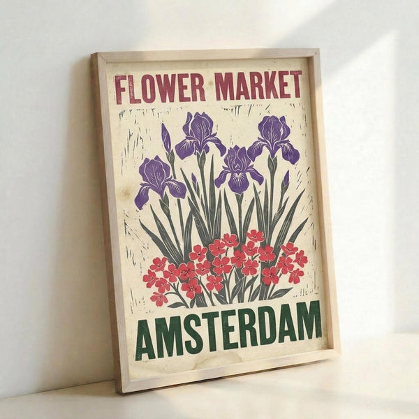 A vintage-style poster in a light wooden frame leaning against a white wall. The cream-colored print features purple irises and red flowers with green stems and leaves in a folk art illustration style. "FLOWER MARKET" appears in burgundy letters at the top, with "AMSTERDAM" in green letters at the bottom. The artwork has a weathered, textured appearance with subtle distressing around the edges.